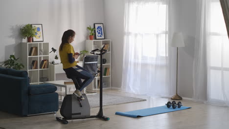 woman-is-sitting-on-stationary-bike-in-apartment-at-weekend-and-browsing-internet-with-smartphone-break-at-morning-workout-of-young-sporty-lady